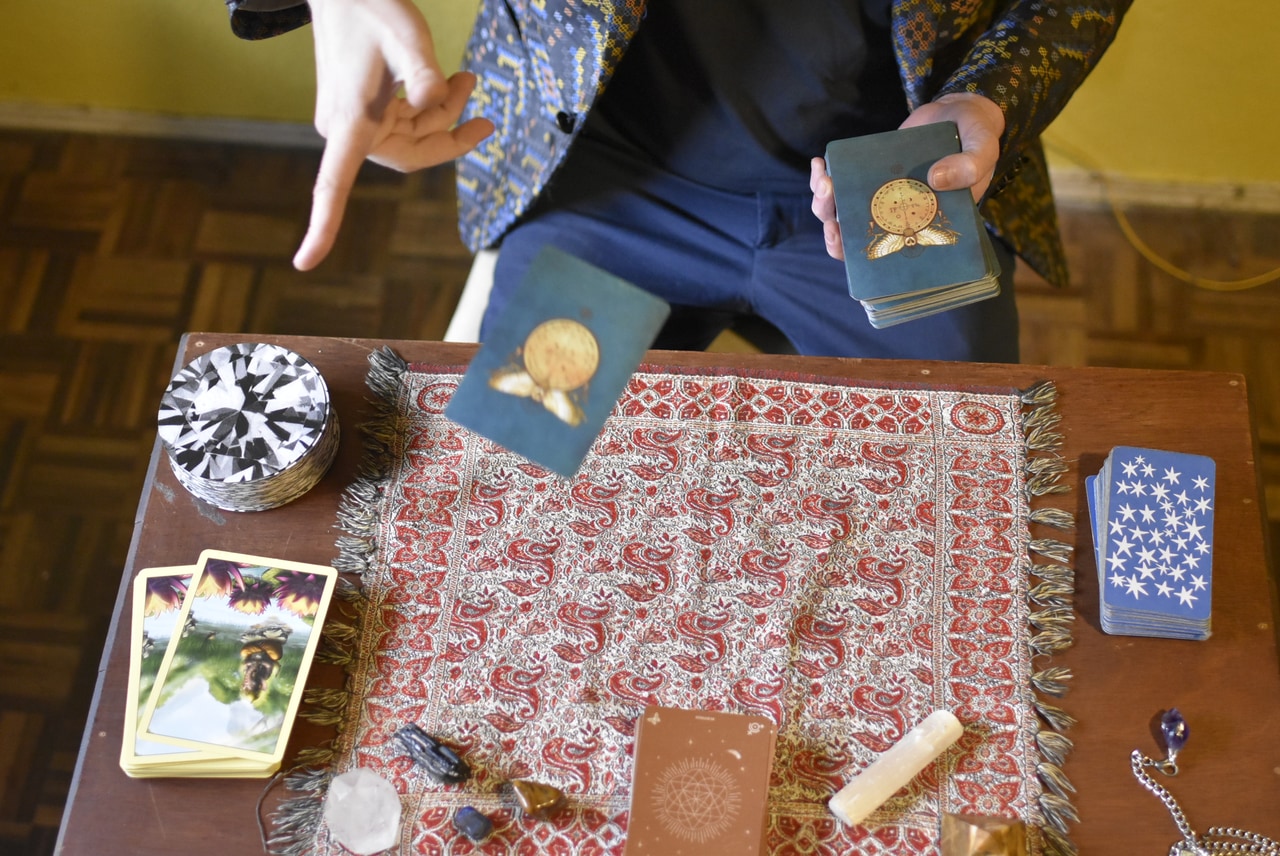 Vista desde arriba: una carta de tarot cae sobre un mantel rojo estampado, entre mazos, cristales y un péndulo.
